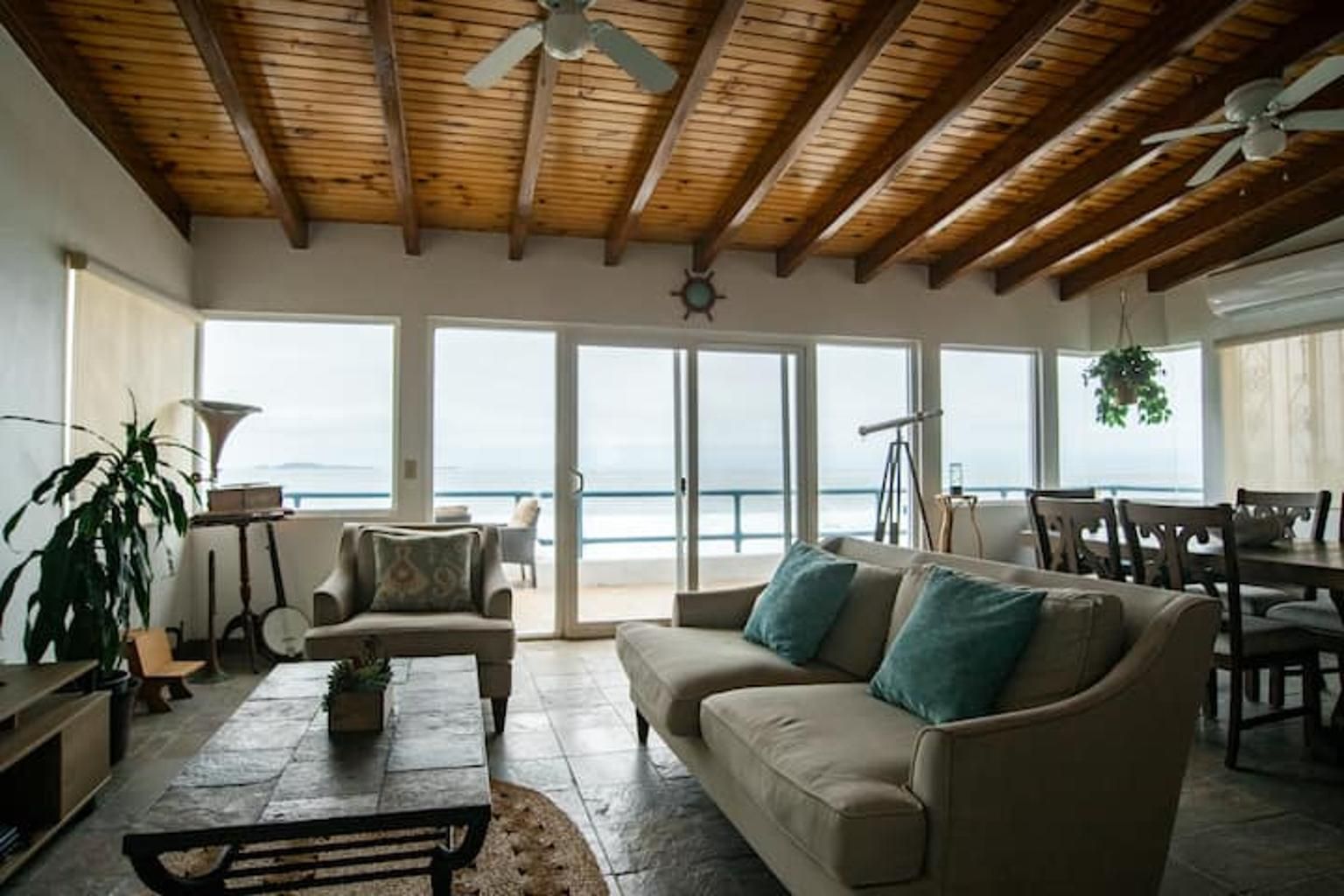 A spacious living area with a wooden ceiling, a sofa with cushions, a coffee table, and a view of the ocean through large sliding doors.