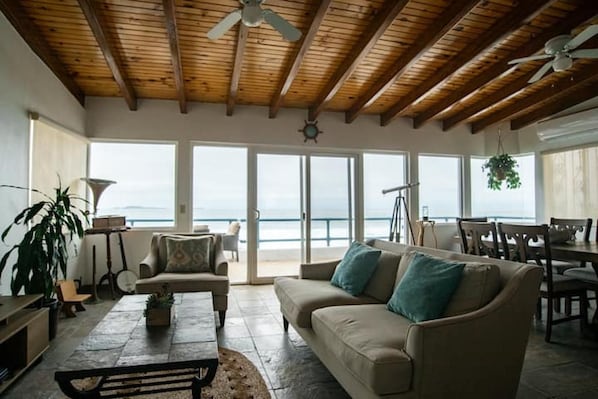 A spacious living area with a wooden ceiling, a sofa with cushions, a coffee table, and a view of the ocean through large sliding doors.