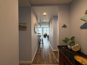A hallway with a wooden floor, a dining area in the background, and a plant on the right side. -  2-bedroom beach condo steps from the beach with amazing sunsets (Virginia Beach)