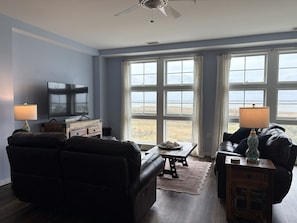 A living room with a flat-screen TV, two sofas, a coffee table, and a view of the outdoors through large windows. -  2-bedroom beach condo steps from the beach with amazing sunsets (Virginia Beach)