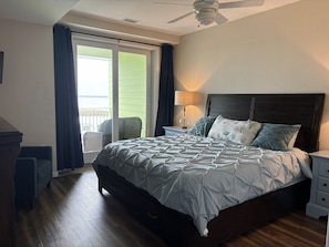 Een slaapkamer met een bed, nachtkastje, lamp, plafondventilator, raam met gordijnen en een stoel.