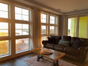 A living room with a brown leather sofa, a wooden coffee table, and large windows with curtains. -  2-bedroom beach condo steps from the beach with amazing sunsets (Virginia Beach)