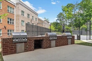 An outdoor barbecue area with three grills, a brick wall, and a fence. - Crimson Tide Condo 3BR w/all kings near Bryant-Denny Stadium | Pool & Jumbotron! (Tuscaloosa)
