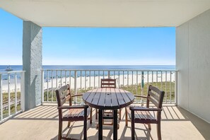 Een balkon met een tafel en stoelen, met uitzicht op een strand met een hek en een heldere lucht.