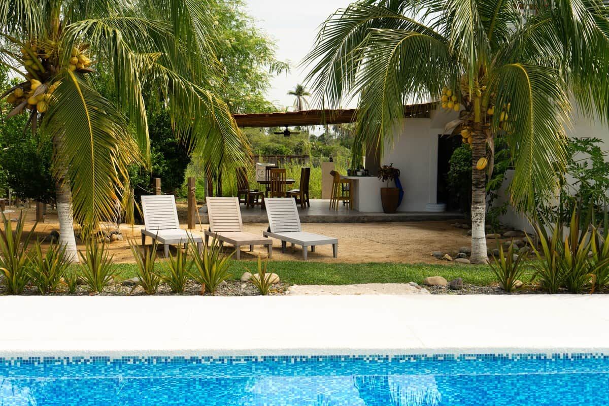 A pool area with lounge chairs, palm trees, and a building with a covered patio.