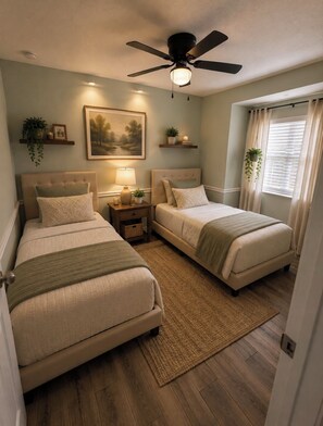 A bedroom with two beds, a ceiling fan, bedside lamps, a painting, and a window with curtains.
