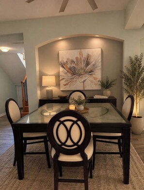 A dining area with a glass-top table, four chairs, a large abstract painting, a potted plant, and a lamp on a side table.