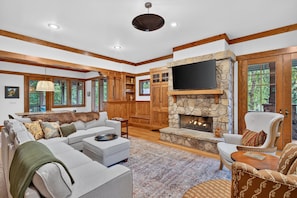 A spacious living room with a stone fireplace, wooden beams, a flat-screen TV, and a large window overlooking greenery. - Day's End Retreat | Pets, Game Room, Fire Pit & Views in Cheshire Village! (Black Mountain)