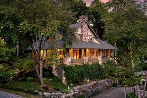 A stone house with a porch, surrounded by trees and a stone wall. - Day's End Retreat | Pets, Game Room, Fire Pit & Views in Cheshire Village! (Black Mountain)