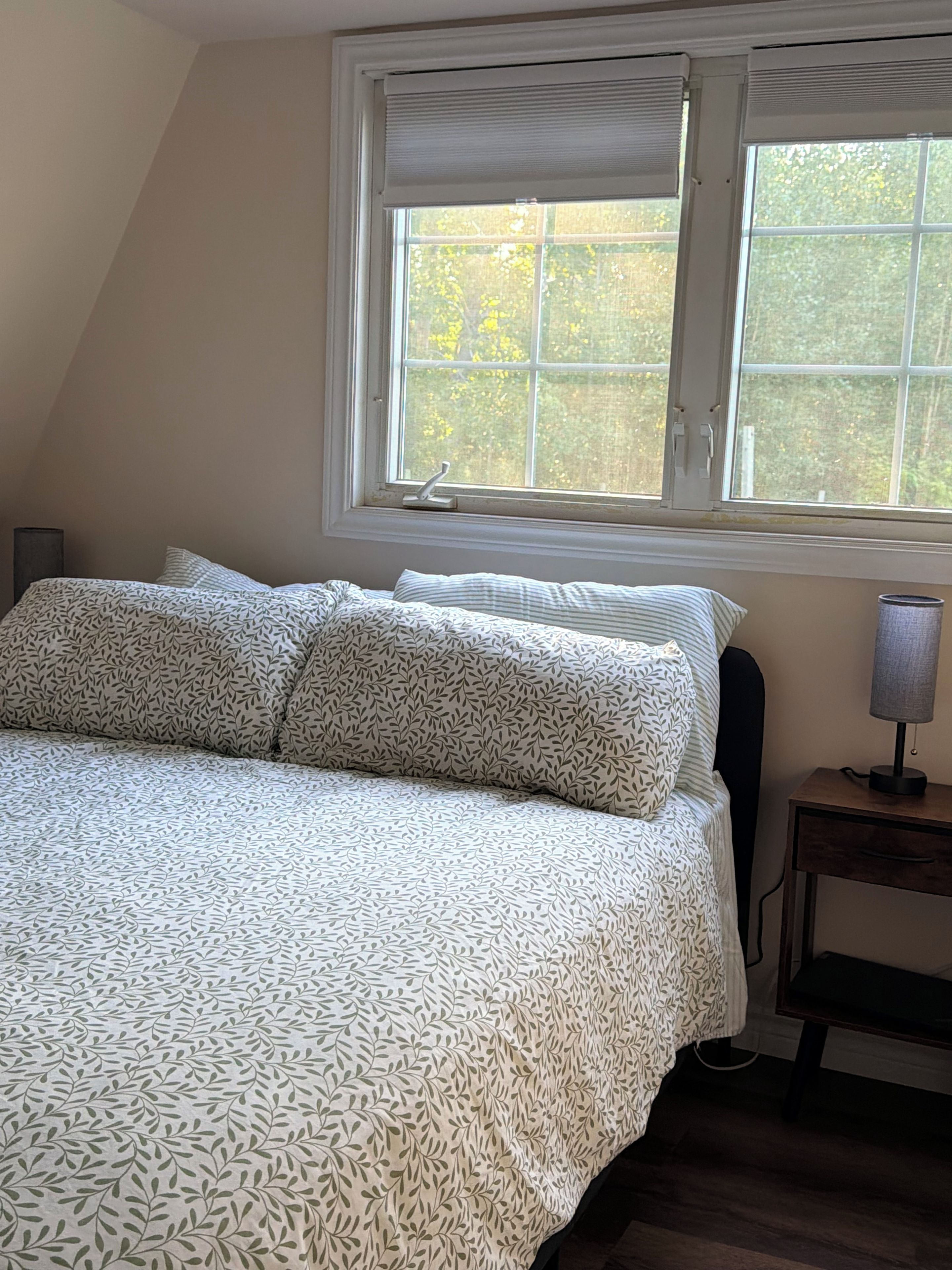 Un dormitorio con cama, almohadas, mesita de noche, lámpara y ventana con persianas.