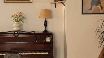 A piano with a chair in front, a lamp, a painting, and a shelf with books and a plant.