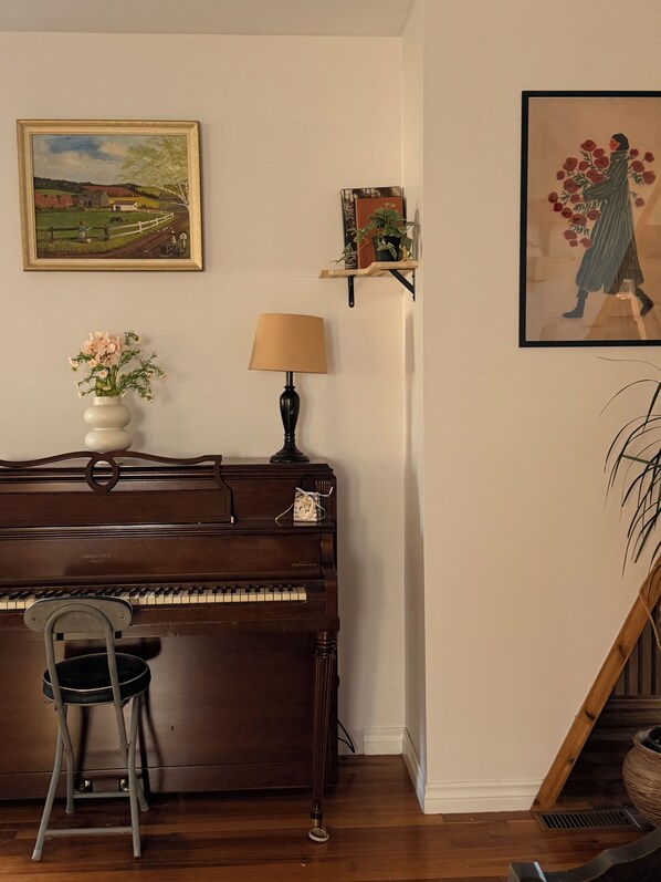 A piano with a chair in front, a lamp, a painting, and a shelf with books and a plant.