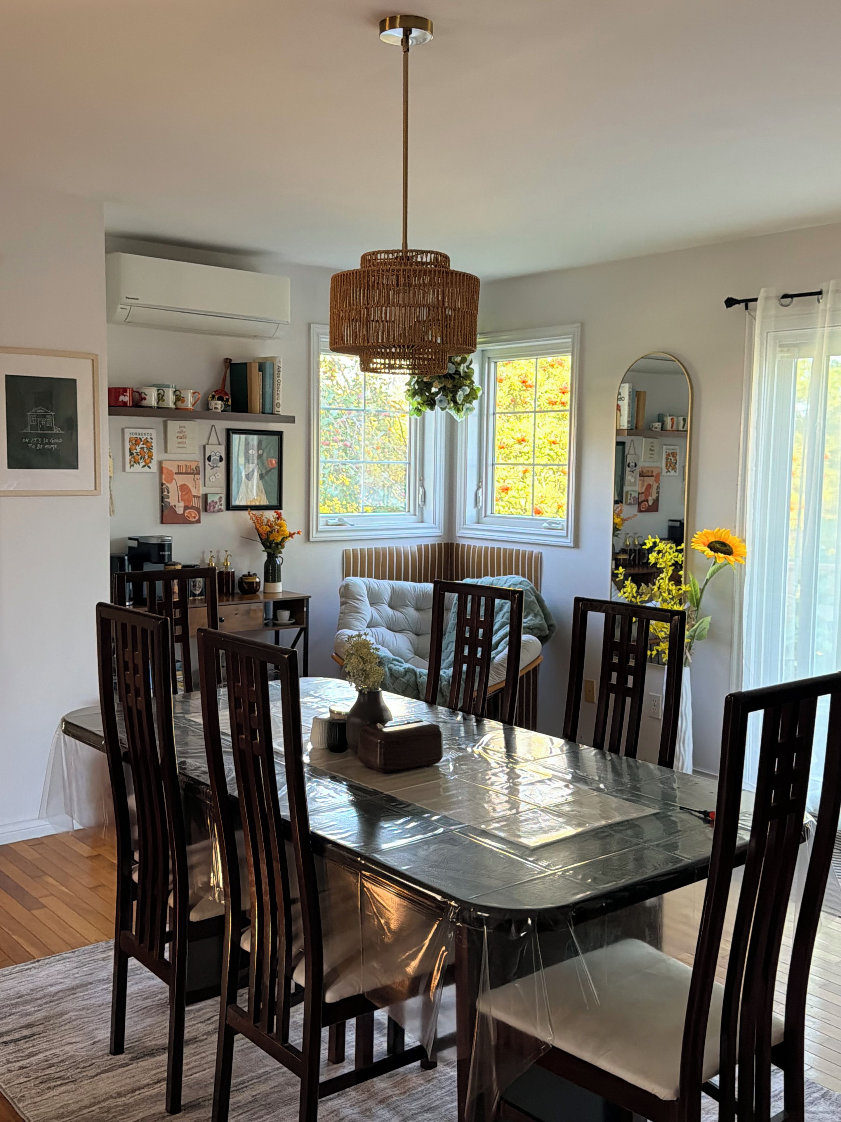 A dining area with a large table, chairs, a hanging light fixture, and a window with a view of trees.