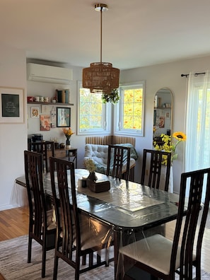 A dining area with a large table, chairs, a hanging light fixture, and a window with a view of trees.