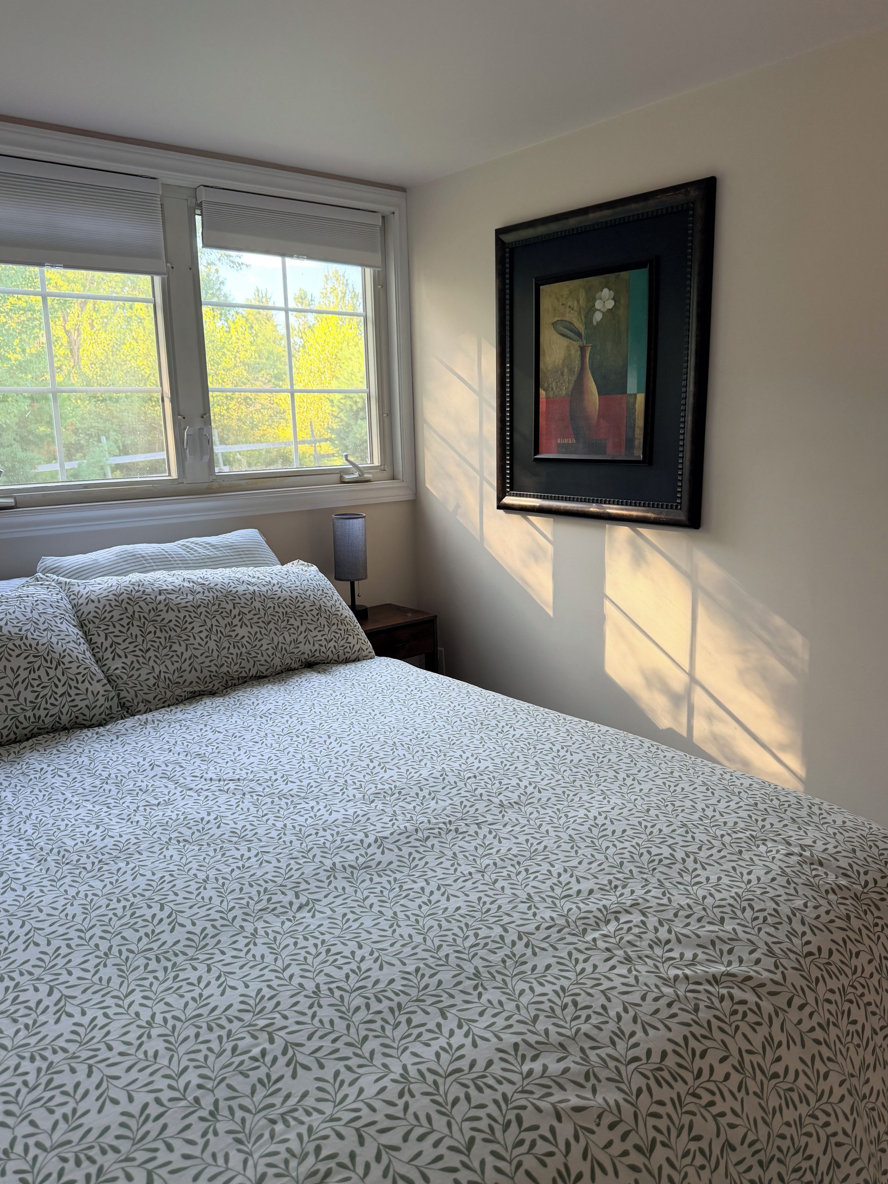 Ein Schlafzimmer mit einem Bett, einem Nachttisch, einer Lampe, einem Fenster mit Jalousien und einem gerahmten Bild an der Wand.