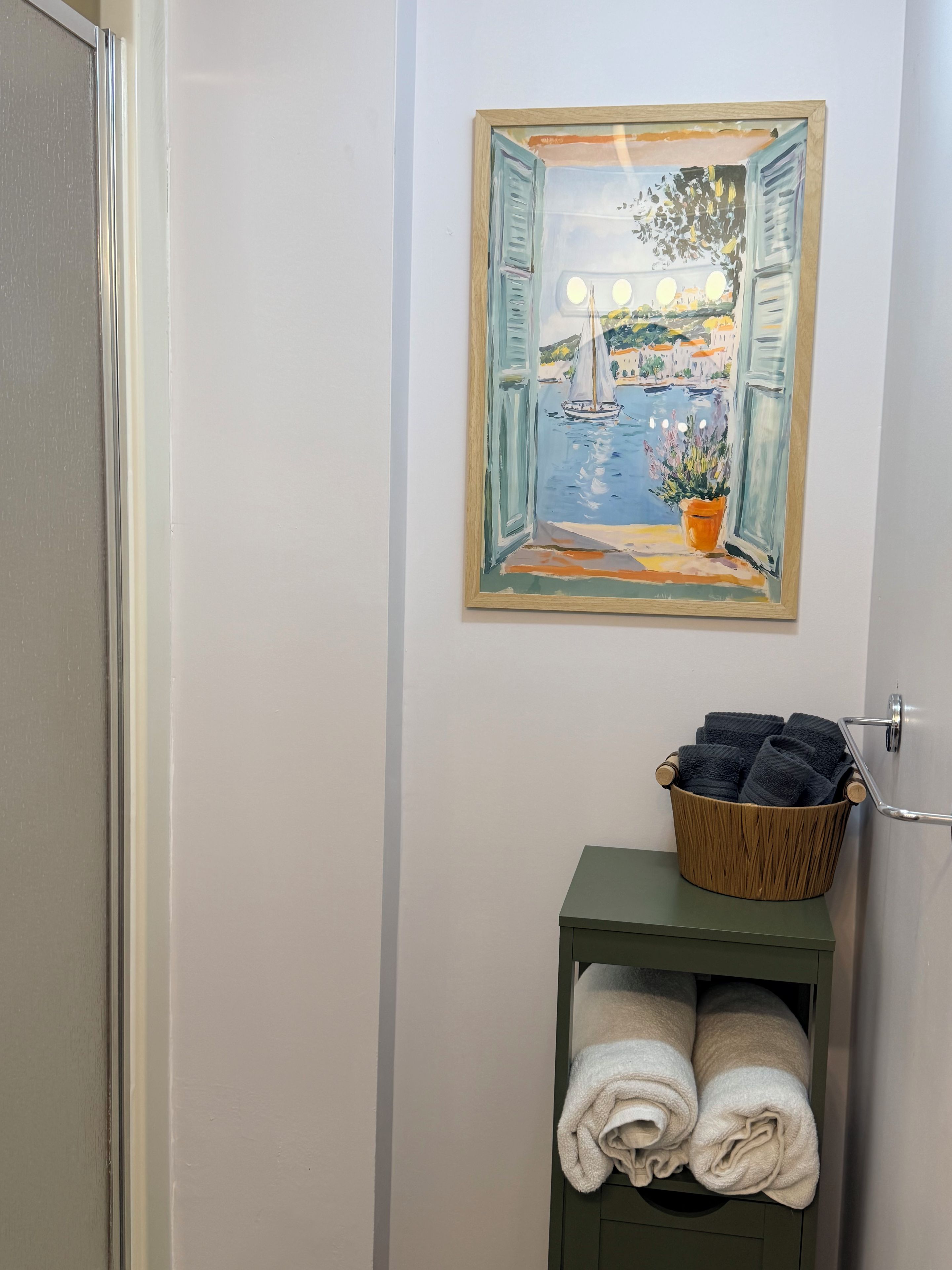 Une salle de bain avec un tableau représentant un paysage côtier, un panier en bois contenant des serviettes et un porte-serviettes vert.