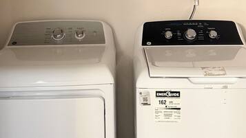 Two front-loading washing machines side by side with a framed sign above them and a basket with folded clothes on the shelf.