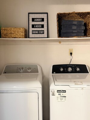 Two front-loading washing machines side by side with a framed sign above them and a basket with folded clothes on the shelf.