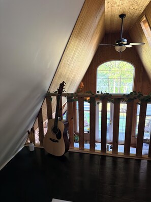 Una habitación con techo inclinado, ventilador de techo, un ventanal con vistas al exterior y una guitarra apoyada en la pared.