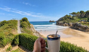 A beach with a clear view of the ocean, a person holding a mug, and a path leading to the beach. - House in Llanes | Directly on the beach | Rebellín (Celorio (Llanes))