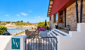 A stone building with a red roof overlooking a beach. There is a patio with a table and chairs, and a sign with the letter 'V'. - House in Llanes | Directly on the beach | Rebellín (Celorio (Llanes))