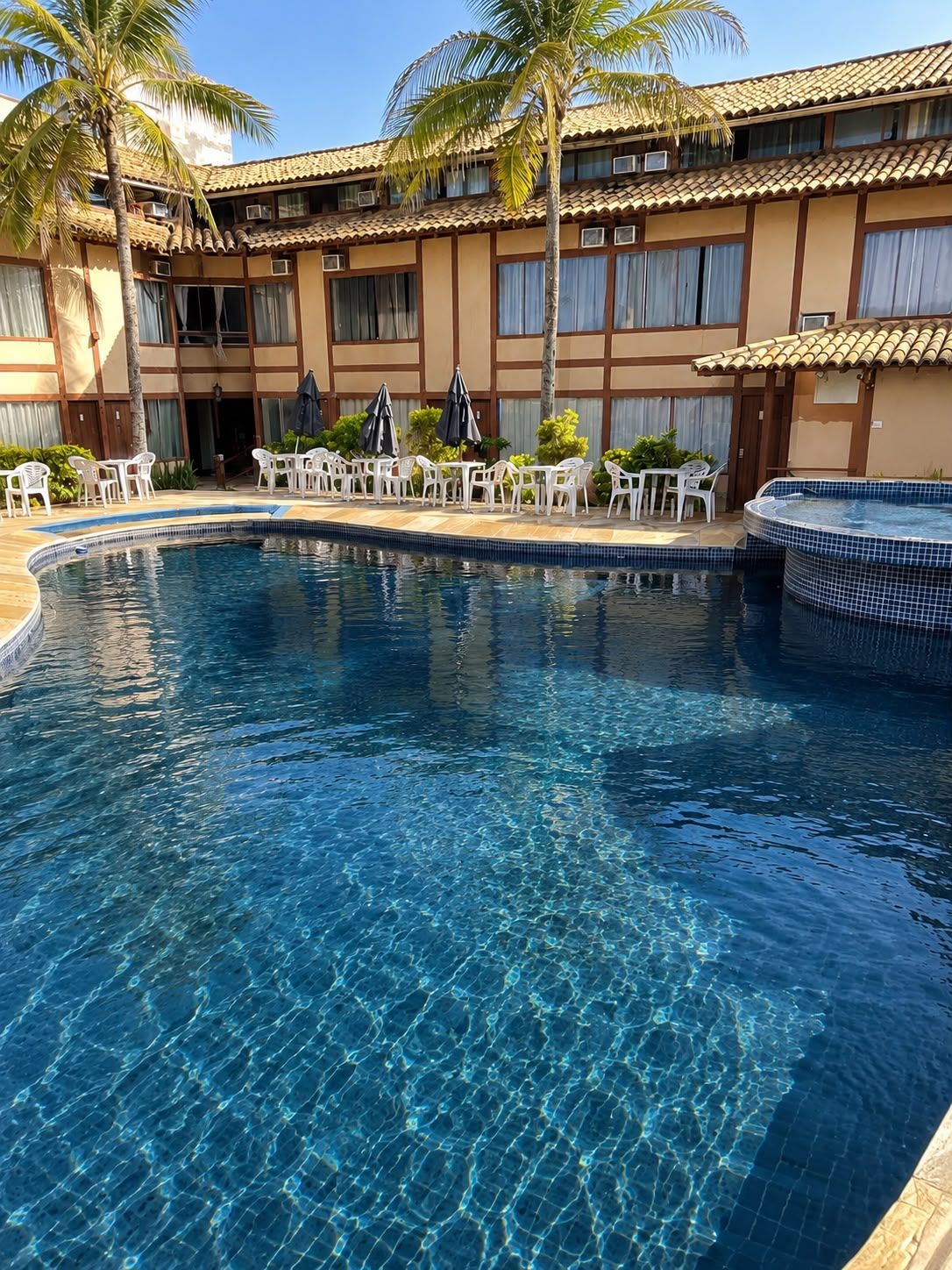 A swimming pool with clear water, surrounded by white chairs and umbrellas, and a building with a tiled roof in the background.