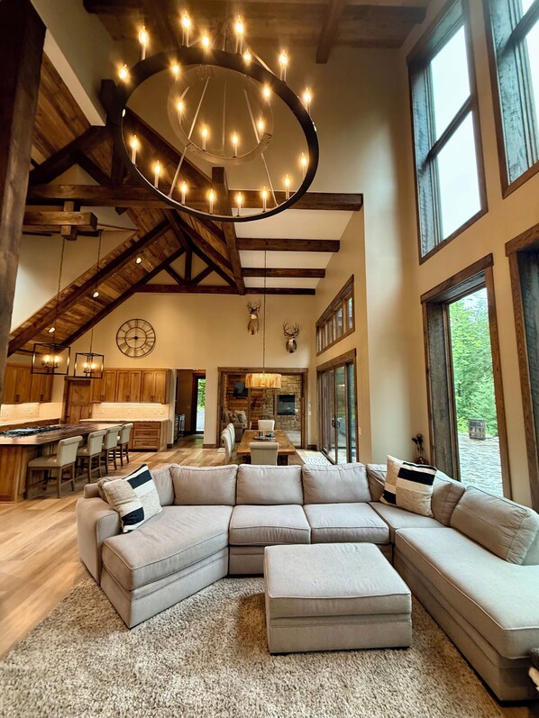 Un vaste espace de vie comprenant un grand canapé d’angle, un plafond en bois avec des poutres apparentes et un luminaire circulaire.