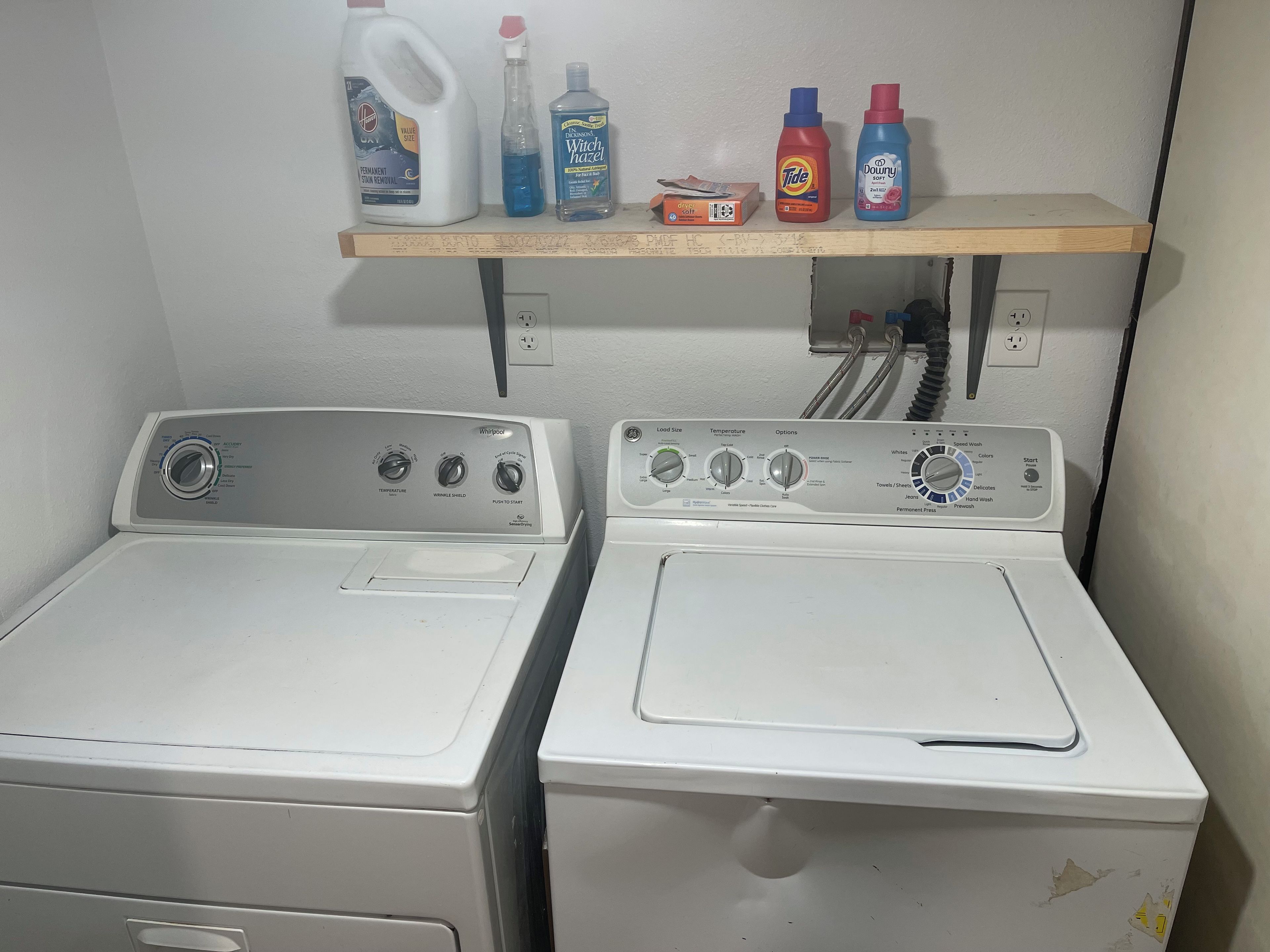 Deux machines à laver à chargement frontal, dotées de panneaux de commande, une étagère au-dessus contenant divers produits de nettoyage, et des prises électriques au mur.