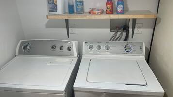 Deux machines à laver à chargement frontal, dotées de panneaux de commande, une étagère au-dessus contenant divers produits de nettoyage, et des prises électriques au mur.