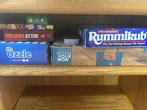 A shelf with various board games, including 'The Uzzle,' 'Solid Wood Four Row,' 'Exploding Kittens,' and 'Rummikub.'