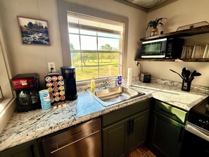 A kitchen with a granite countertop, stainless steel appliances, a window with a view of a green landscape, and a shelf with kitchen utensils. - Serene 1-bedroom tiny house in charming Strawberry, steps away from the creek (Strawberry)