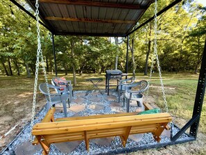 A covered outdoor seating area with a swing, a bench, and a grill table.