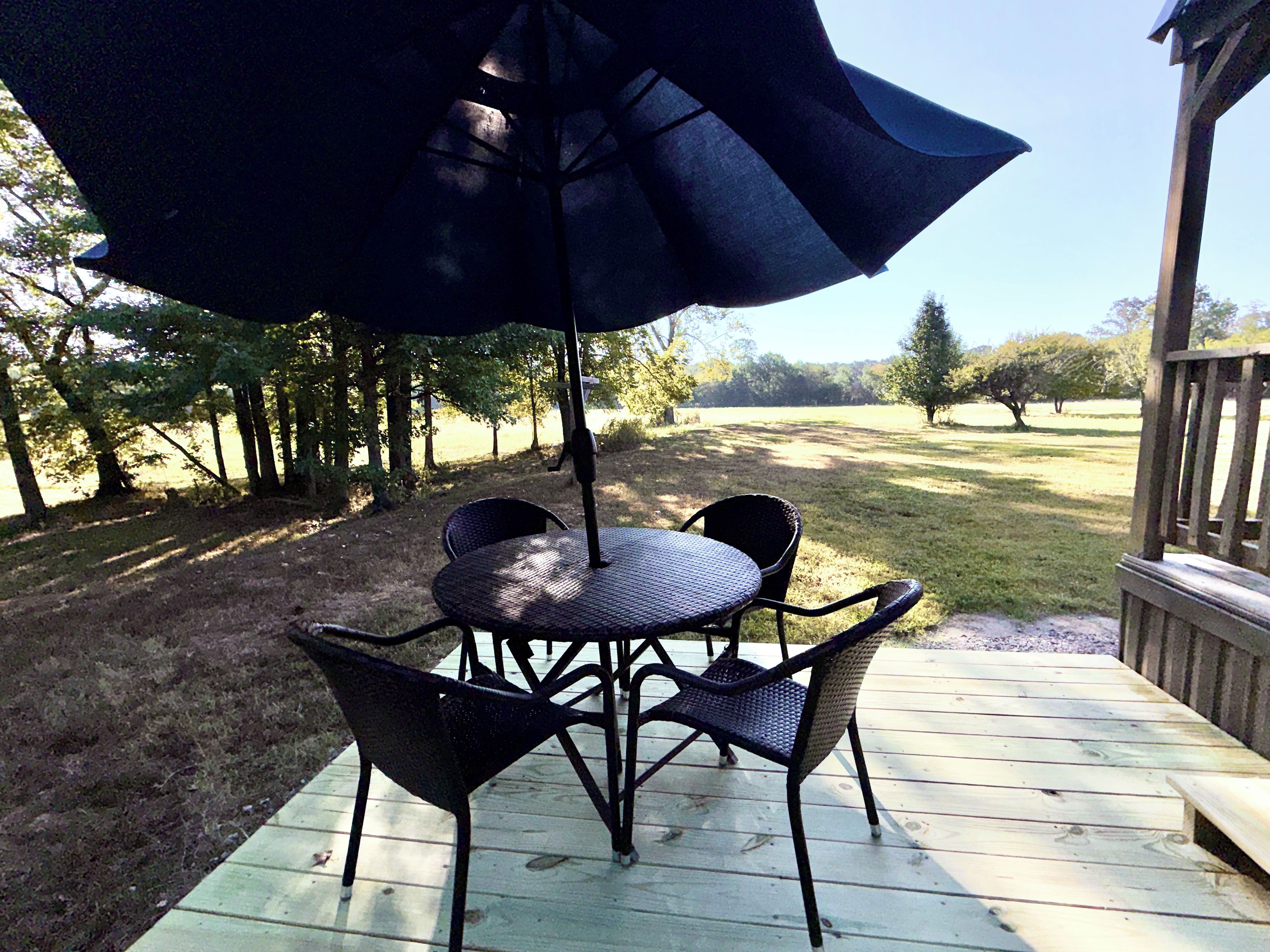 A wooden deck with a round table and four chairs, an umbrella, and a view of a grassy area with trees.