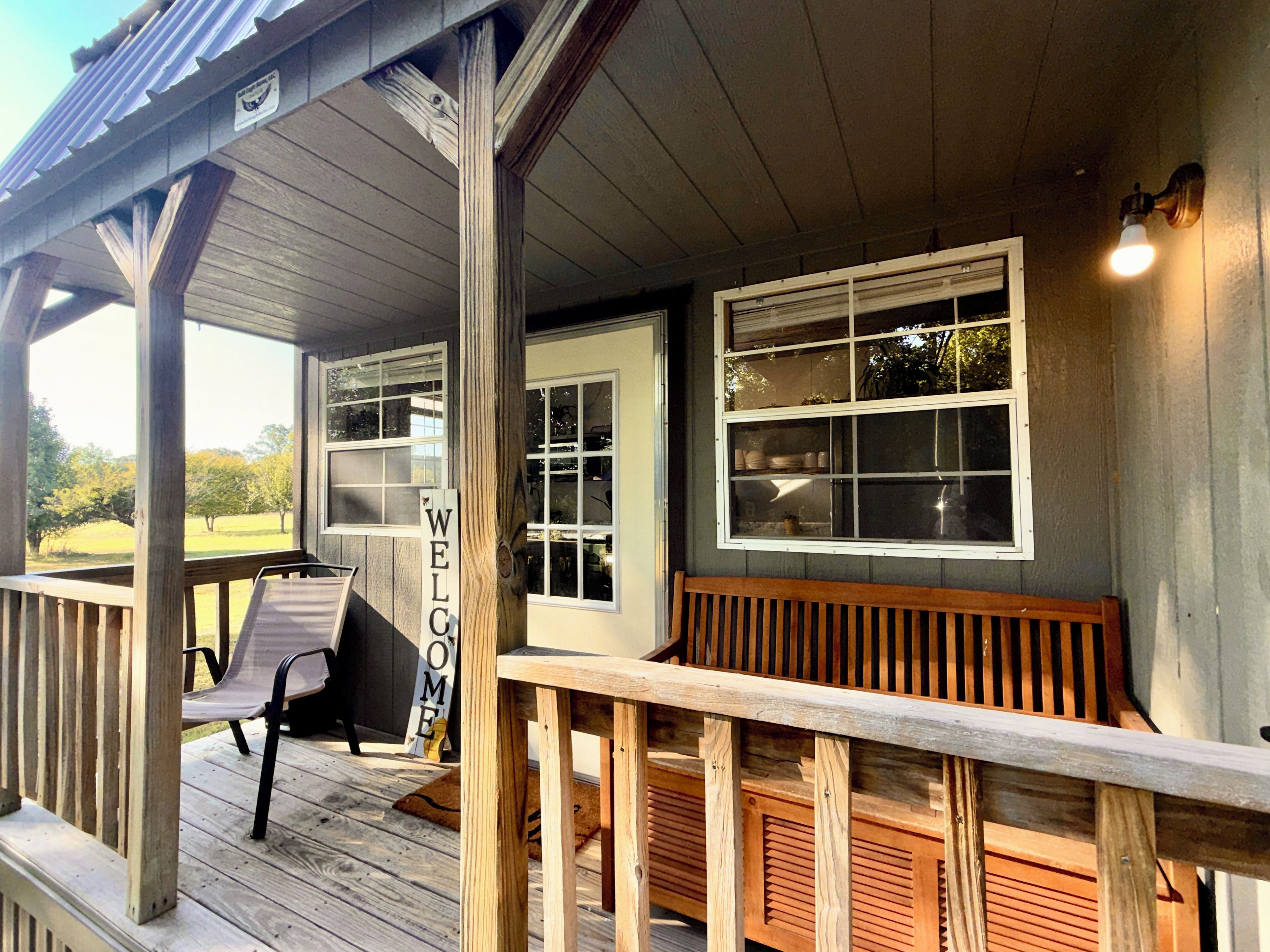 A wooden porch with a bench, a welcome sign, and a window with a view of trees.