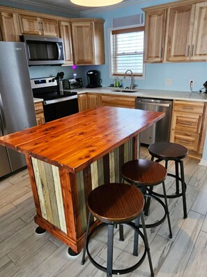 A kitchen with wooden cabinets, a stainless steel refrigerator, a microwave, a coffee maker, and a wooden island with stools. - Bayfront Retreat for Two - Pool + Gulf Nearby (Fort Walton Beach)