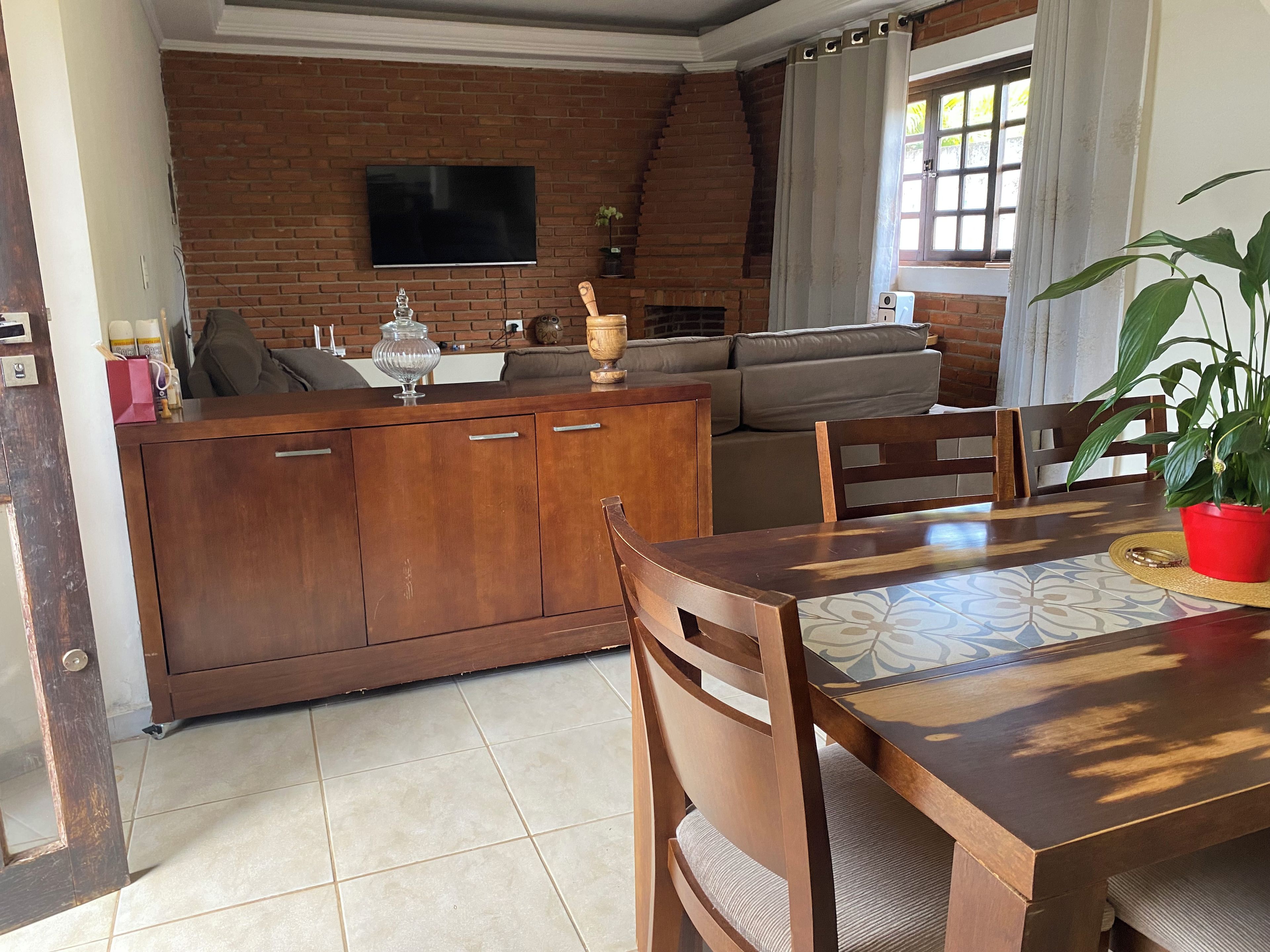 Un coin repas avec une table et des chaises en bois, un mur en briques, un téléviseur fixé au-dessus d’une cheminée et une fenêtre avec des rideaux.