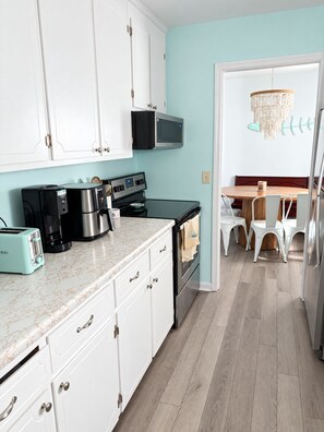 A kitchen with white cabinets, a microwave, a coffee maker, a toaster, and a wooden dining table with chairs. - Peaceful 3-bedroom house with a Heated Pool! (Oak Island)