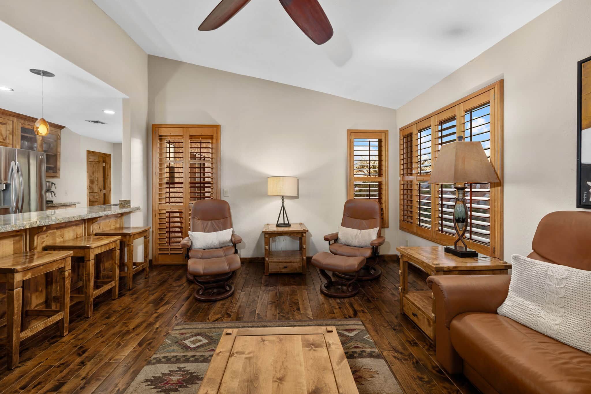 A spacious living room with a wooden floor, leather chairs, a sofa, a dining area with a wooden table, and a kitchen with wooden cabinets.