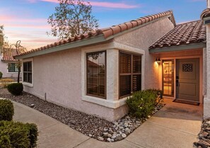 A single-story house with a tiled roof, a front door with a decorative glass panel, and a small garden area with rocks and shrubs. - The Hilltop Lookout | Catalina Mountain Views (Tucson)