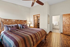Ein Schlafzimmer mit einem hölzernen Kopfteil, einem großen Bett mit einer gestreiften Bettdecke, einem Deckenventilator und einer Holztür, die ins Bad führt.