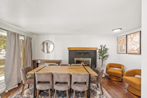 A modern dining room with a large wooden table, six chairs, a fireplace, and a mirror on the wall.