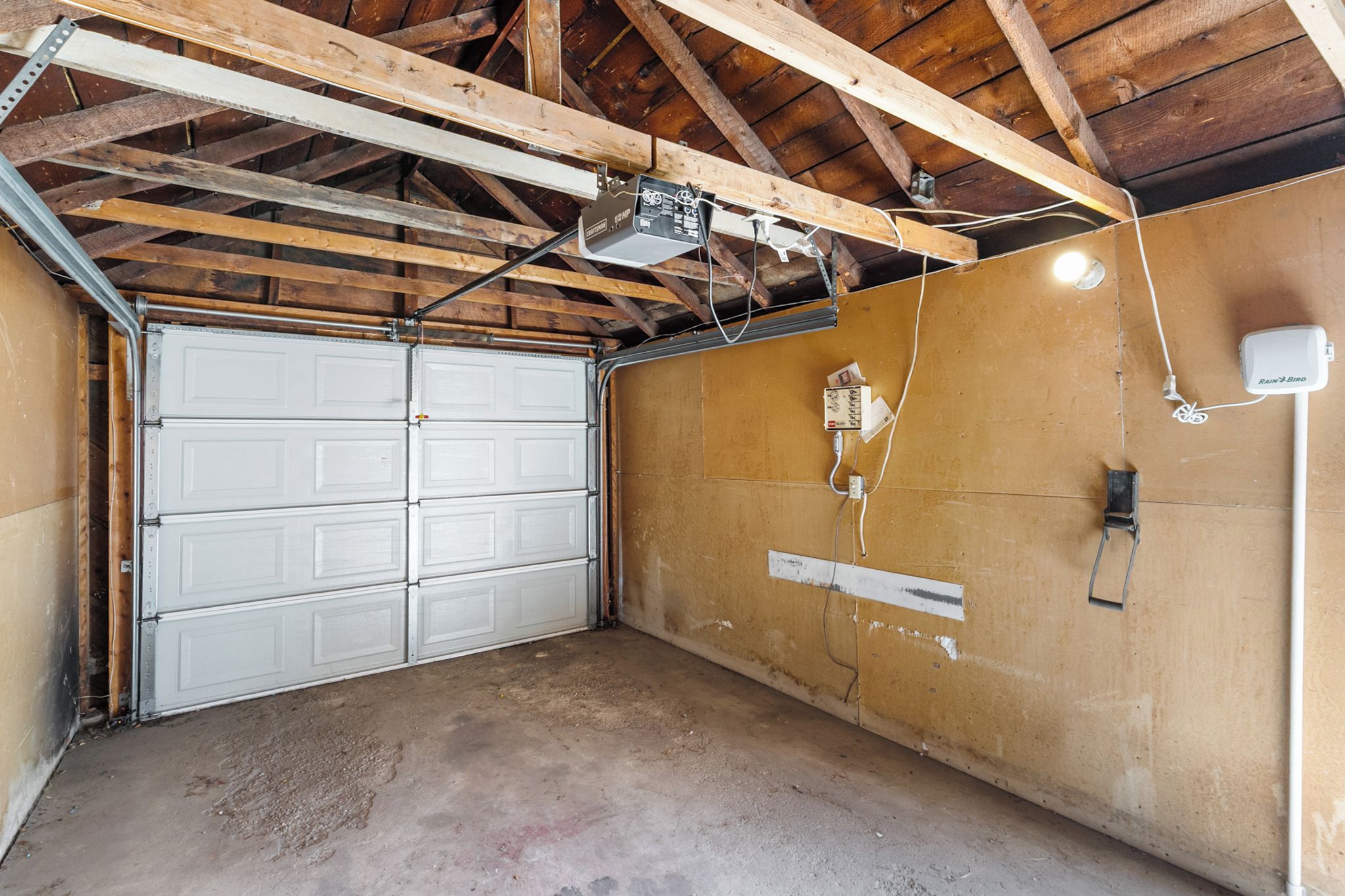 Un garage avec une porte de garage blanche, des poutres en bois et un sol en béton.
