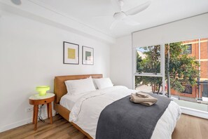 Ein Schlafzimmer mit Bett, Nachttisch, Lampe, gerahmten Bildern an der Wand und einem Fenster mit Blick auf ein Gebäude und Bäume.
