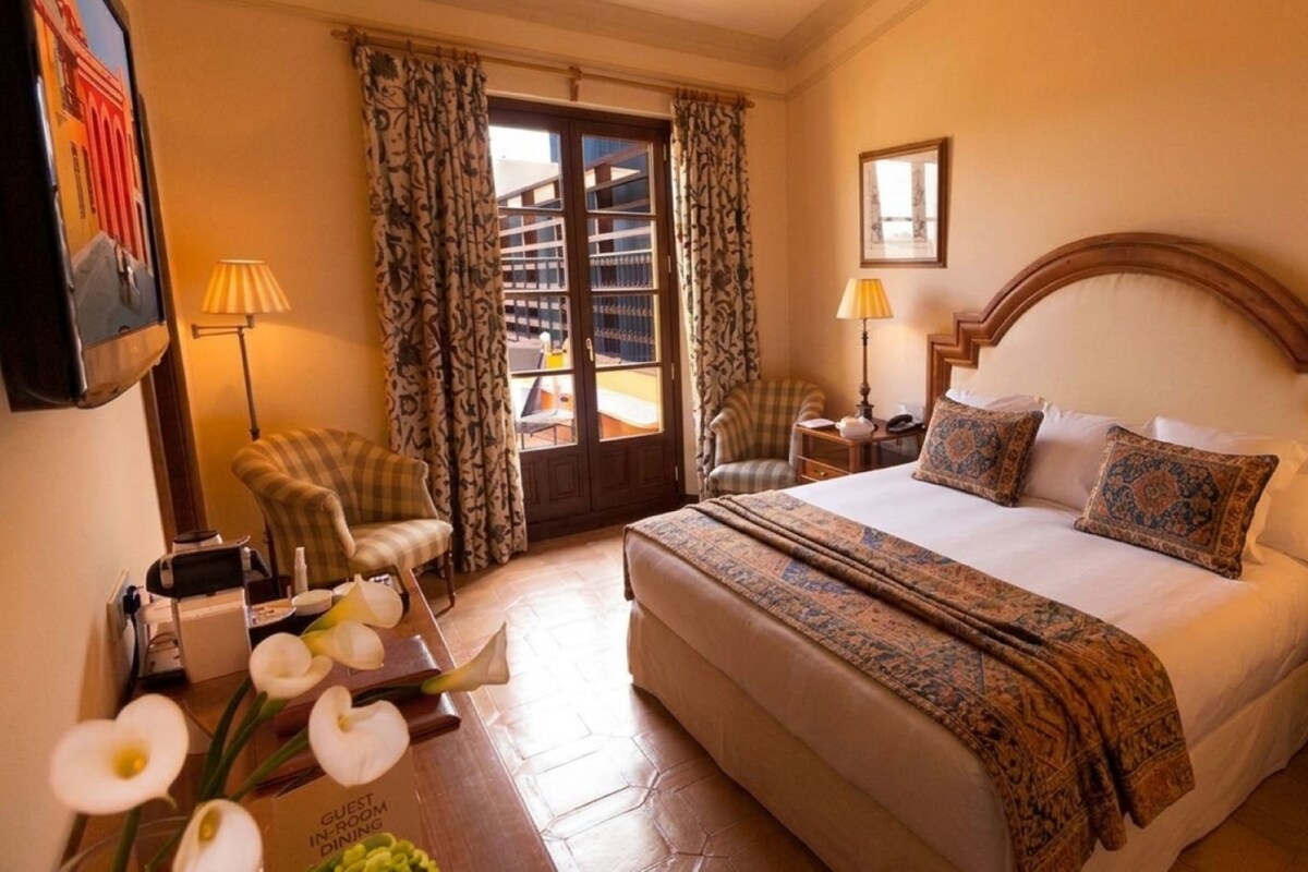 Habitación de hotel con una cama grande, una silla, un escritorio, una lámpara y una ventana con cortinas.