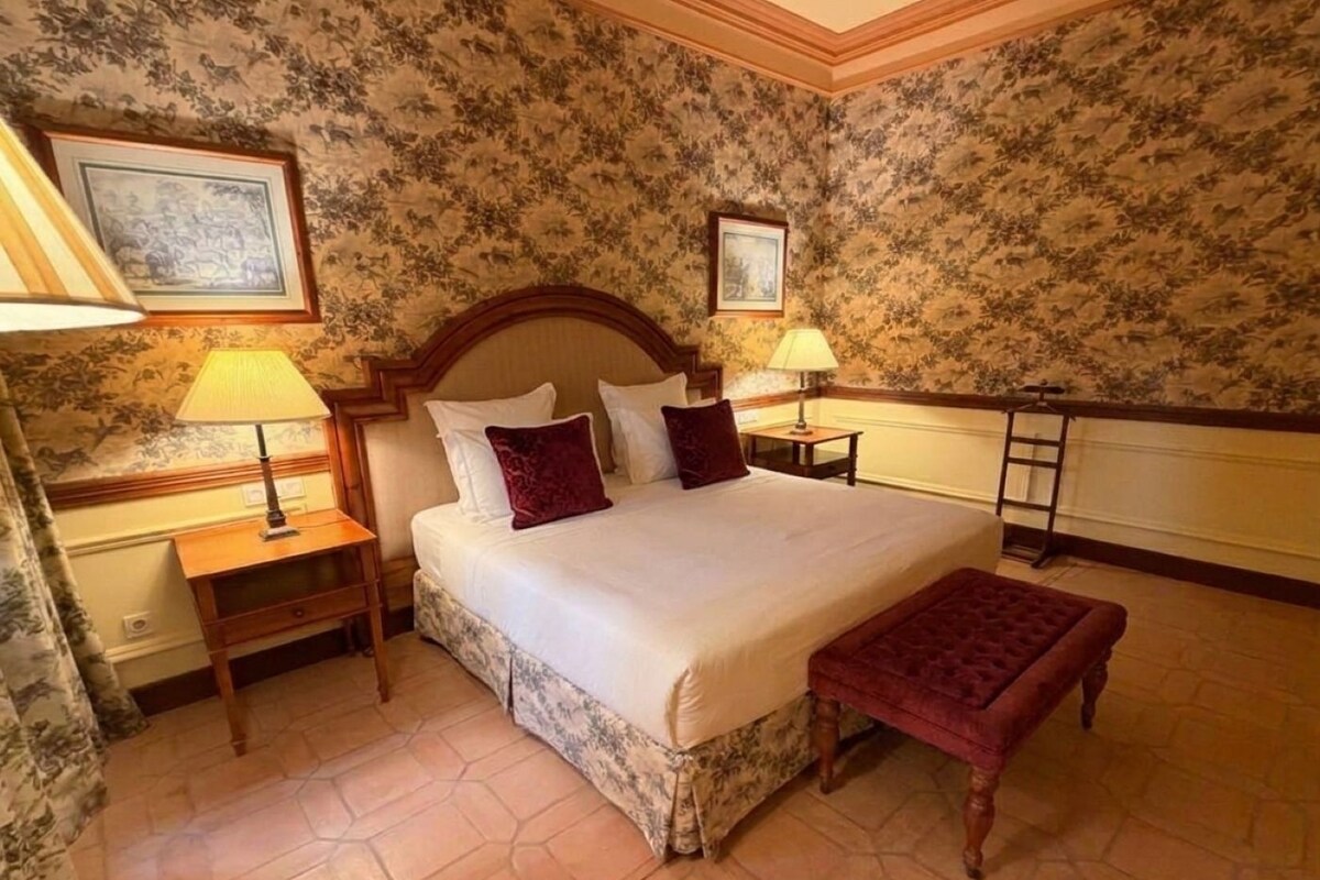 Una habitación de hotel con una cama grande, dos mesitas de noche con lámparas, un cabecero de madera, papel tapiz floral y un banco.