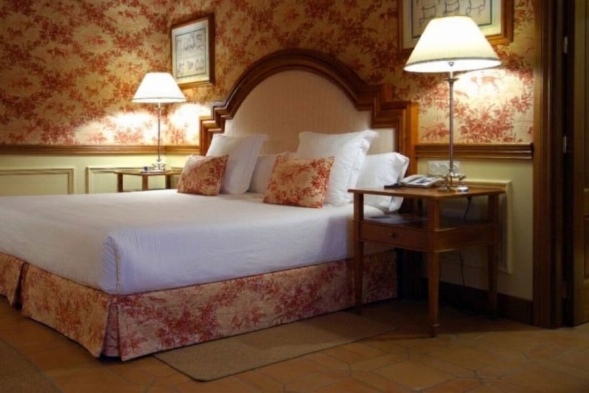 Una habitación de hotel con una cama grande, dos mesitas de noche y un buró con una lámpara.