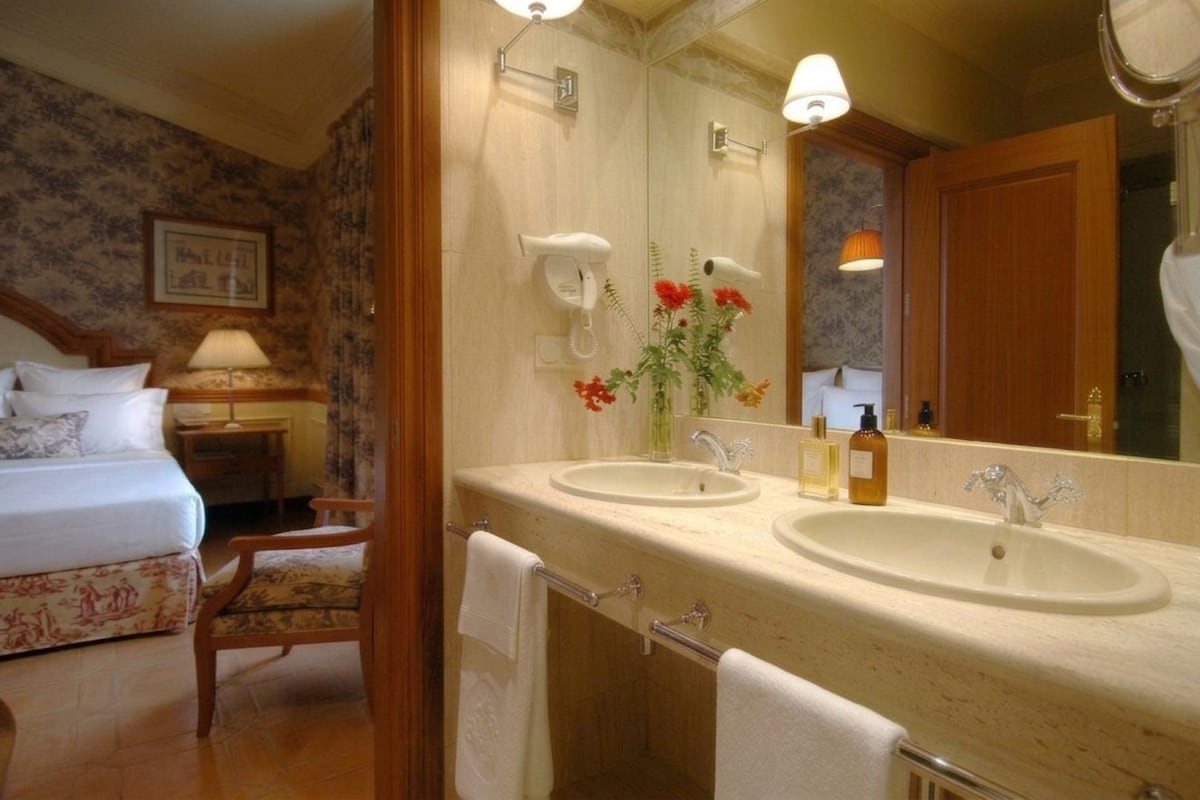 Habitación de hotel con cama, dos lavamanos, espejo, silla y mesita de noche con lámpara.