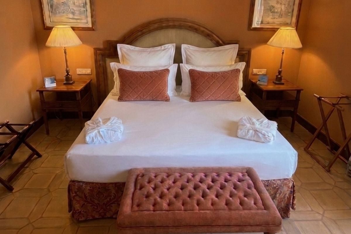 Una habitación de hotel con una cama grande, dos lámparas de noche, un cabecero y un banco.