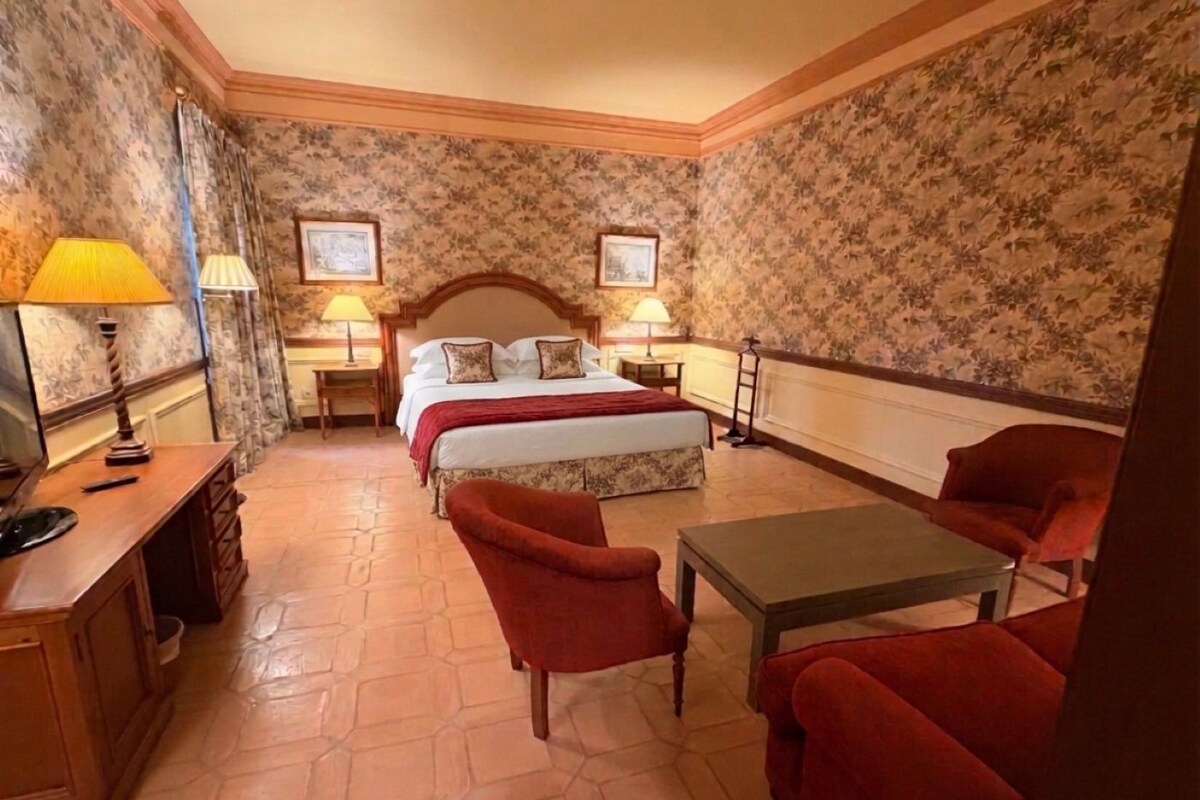 Habitación de hotel con cama, dos mesitas de noche con lámparas, un escritorio con teléfono, dos sillones rojos y una mesita.