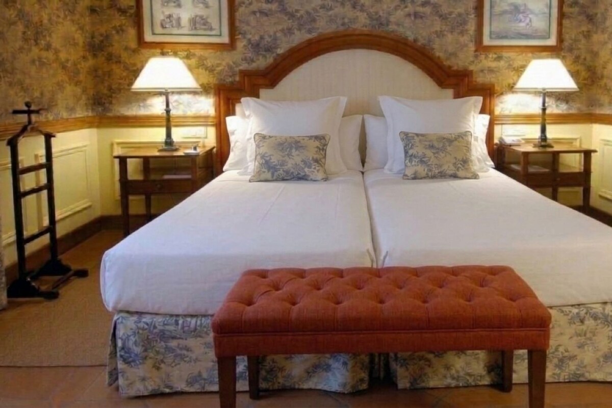 Una cama doble con cabecera de madera, dos mesitas de noche con lámparas y un banco estampado con motivos florales.